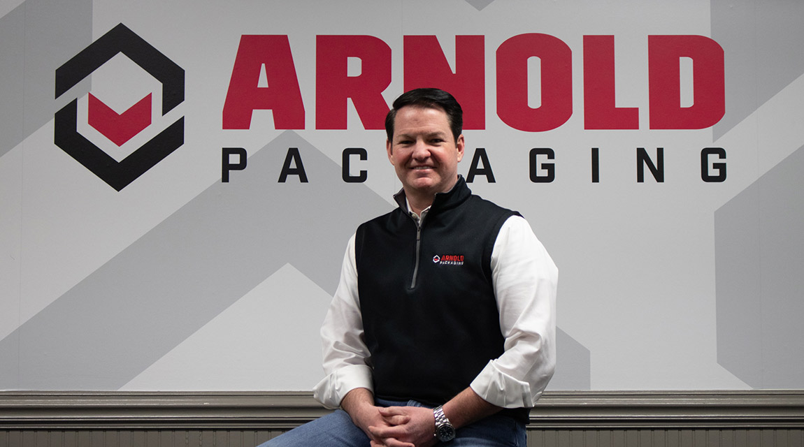 President and CEO Mick Arnold sits on a desk in front of company logo with a jovial look on his face.